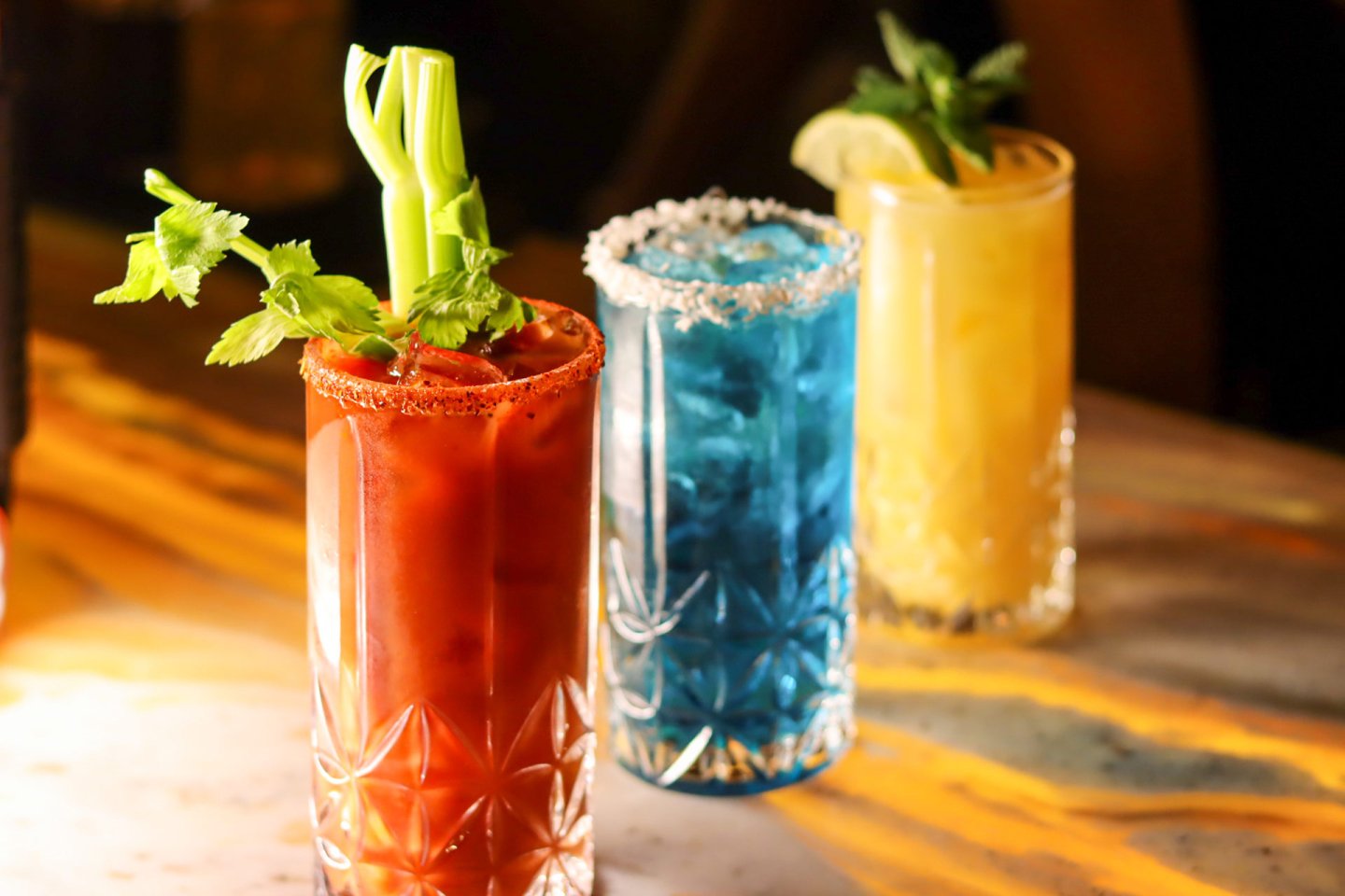 Three colorful cocktails in tall glasses: a red drink with celery and chili rim, a blue drink with a salted rim, and a yellow drink with mint and lime garnish, all on a sunlit marble surface.