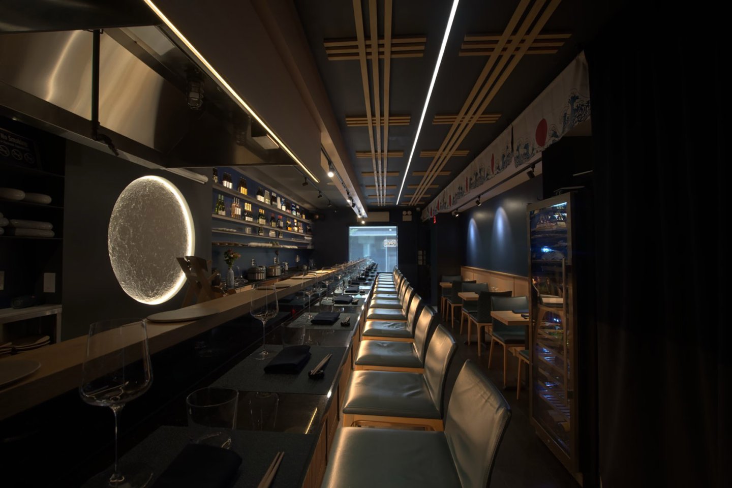 Dimly lit modern sushi bar with a long counter, barstools, and a glowing circular light on the wall. Tables line the opposite side, and shelves with bottles and plates are visible in the background.