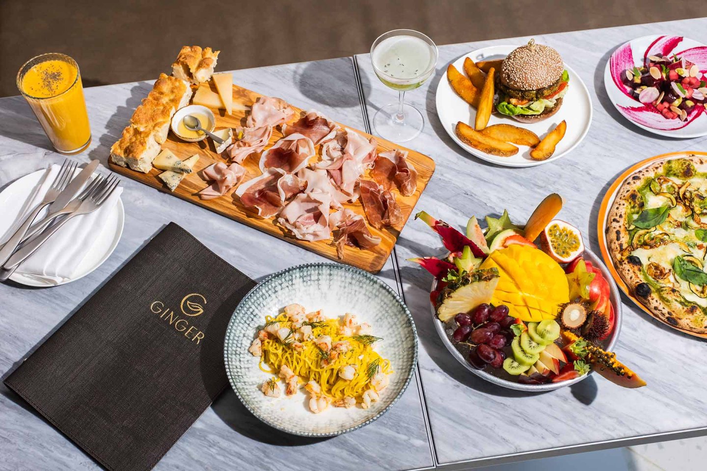 An overhead view of a table with assorted foods: charcuterie board, burger with wedges, salad, fruit bowl, pizza, a cocktail, orange juice, utensils, and a restaurant menu labeled "Ginger.