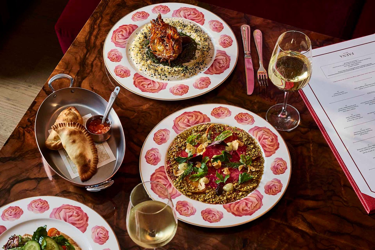 Three plates of gourmet food and a dish of bread are arranged on a wooden table with two glasses of white wine, cutlery, and a restaurant menu visible in the background.