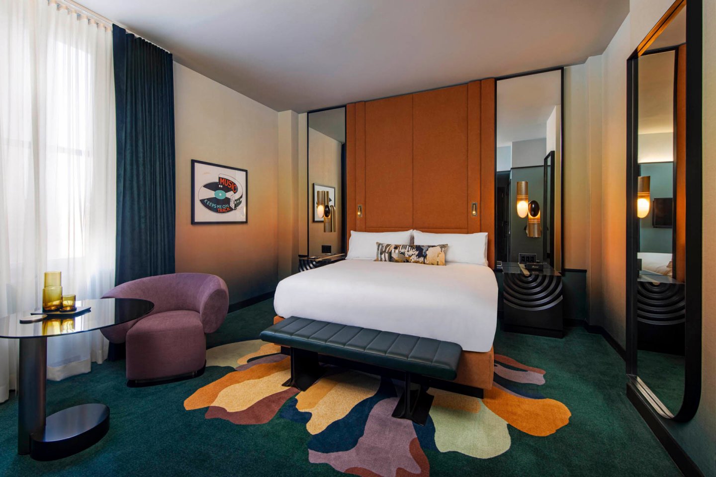 Modern hotel room with a large bed, brown upholstered headboard, colorful abstract rug, purple chair, round glass table, decorative lighting, and tall windows with sheer white curtains and dark drapes.