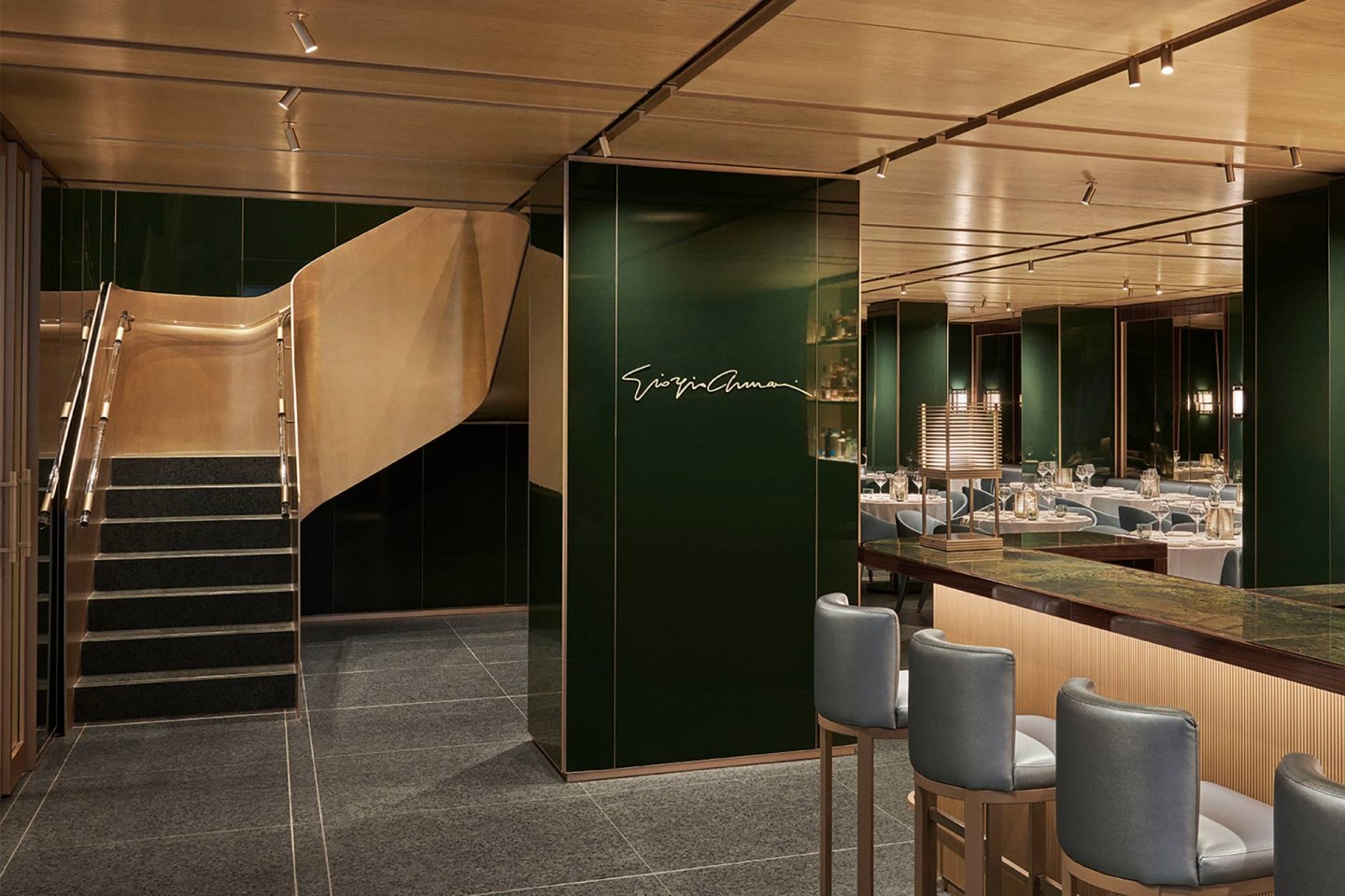 Upscale restaurant interior with green walls, a wooden ceiling, grey tiled floors, blue cushioned bar stools, neatly set tables, and a staircase with wooden handrails. Elegant lighting creates a warm, inviting atmosphere.