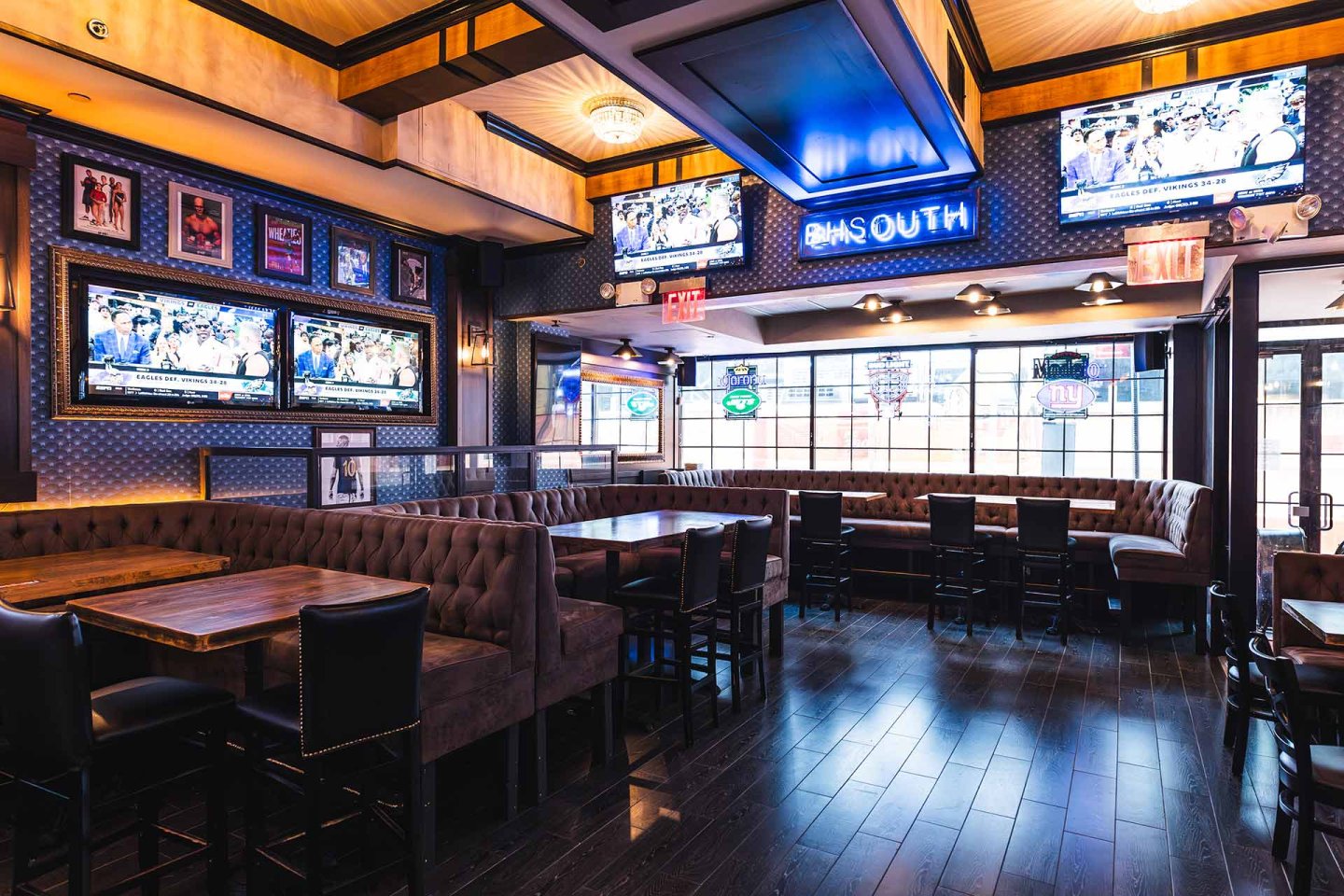 A spacious sports bar with wooden tables, brown cushioned booths, multiple flat-screen TVs on the walls, framed sports memorabilia, and a neon "DMS South" sign above the seating area.