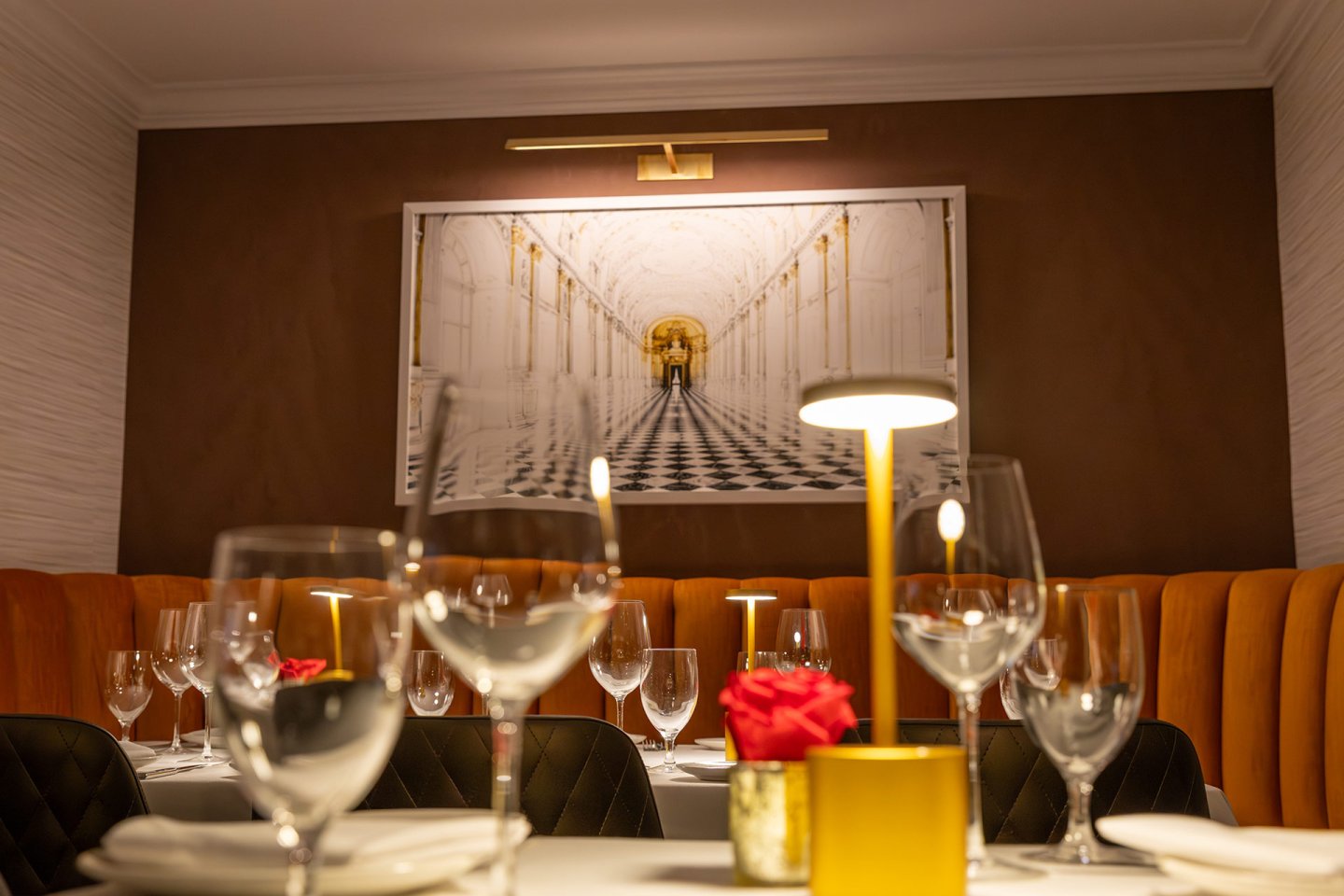 A cozy restaurant table is set with wine glasses, plates, and red rose centerpieces. Warm yellow lighting and an orange cushioned bench create an inviting atmosphere. A framed hallway photograph hangs on the wall behind.