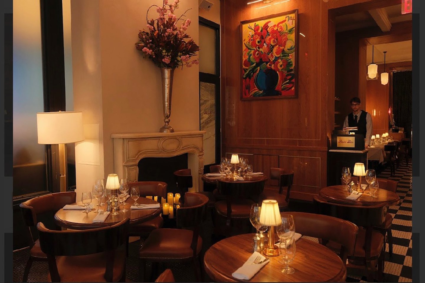 A cozy, dimly lit restaurant with round wooden tables, brown chairs, table lamps, a large floral arrangement on a mantle, and a colorful flower painting on the wall. A host stands by a podium near the entrance.