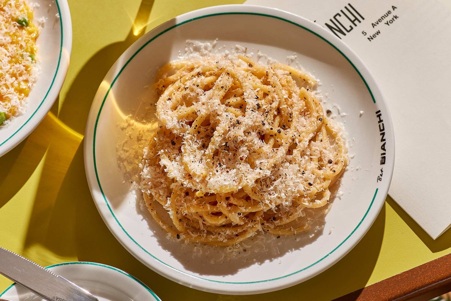 A plate of spaghetti topped with grated cheese and cracked black pepper sits on a table next to a menu and a knife, under bright lighting.