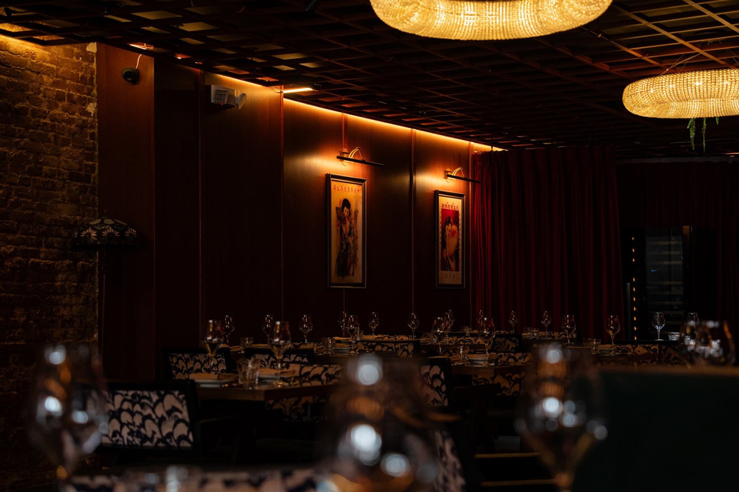 A dimly lit restaurant with elegant chandeliers, red curtains, framed artwork on wood-paneled walls, and tables set with wine glasses and patterned chairs.