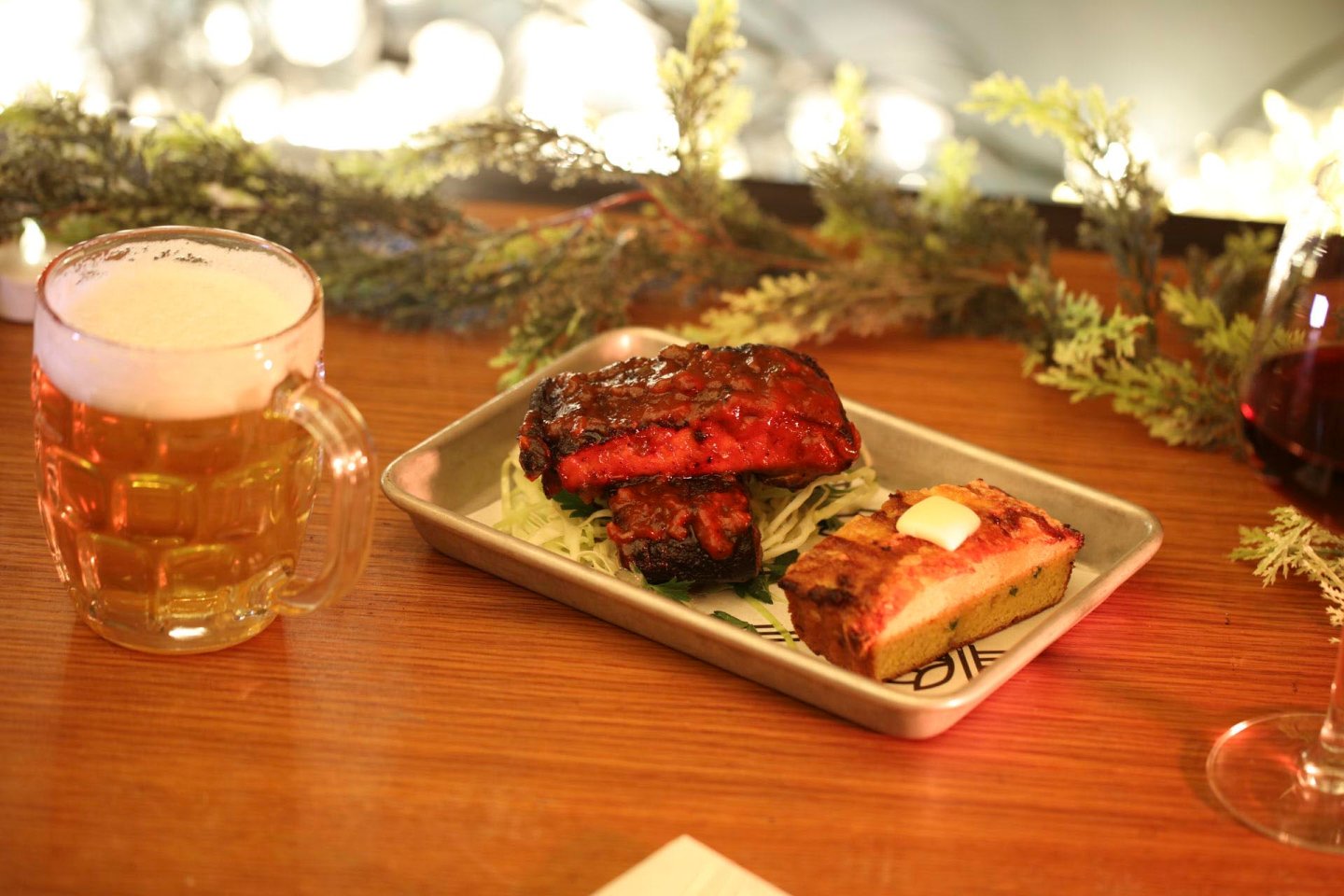 A tray with glazed ribs on lettuce and a slice of cornbread with butter sits on a wooden table. A glass mug of beer is on the left, and a glass of red wine is on the right. Greenery decorates the backdrop.