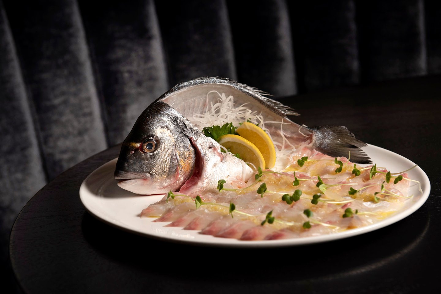 A white plate displays thinly sliced raw fish arranged in a fan, with the fish's head and tail intact, garnished with lemon slices, microgreens, and shredded daikon, set against a dark background.