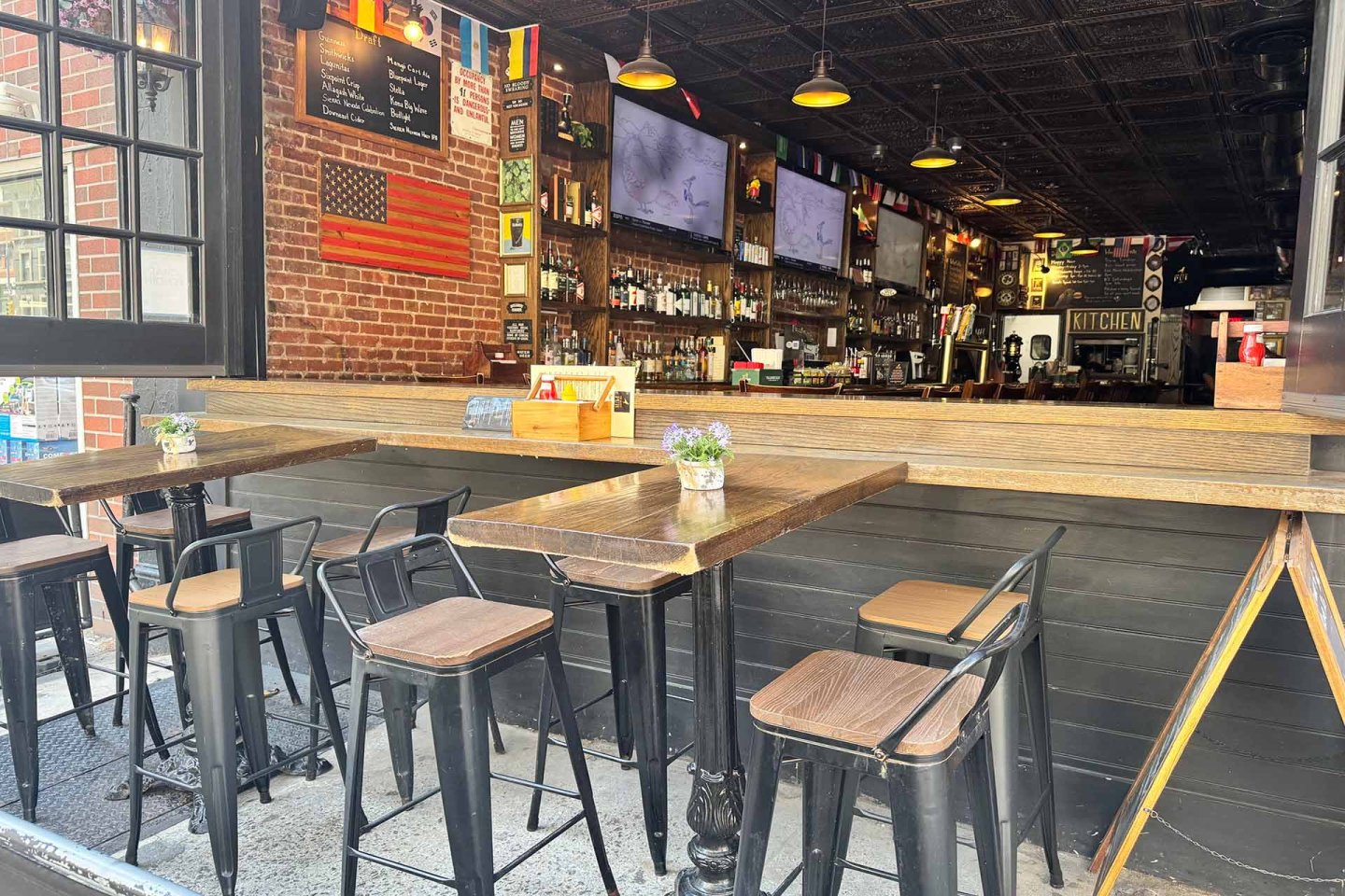 Open-air bar with high wooden tables and metal chairs, flower pots on tables, exposed brick walls, American flag, multiple TVs, and a well-stocked bar inside. Warm lights create a cozy, inviting atmosphere.