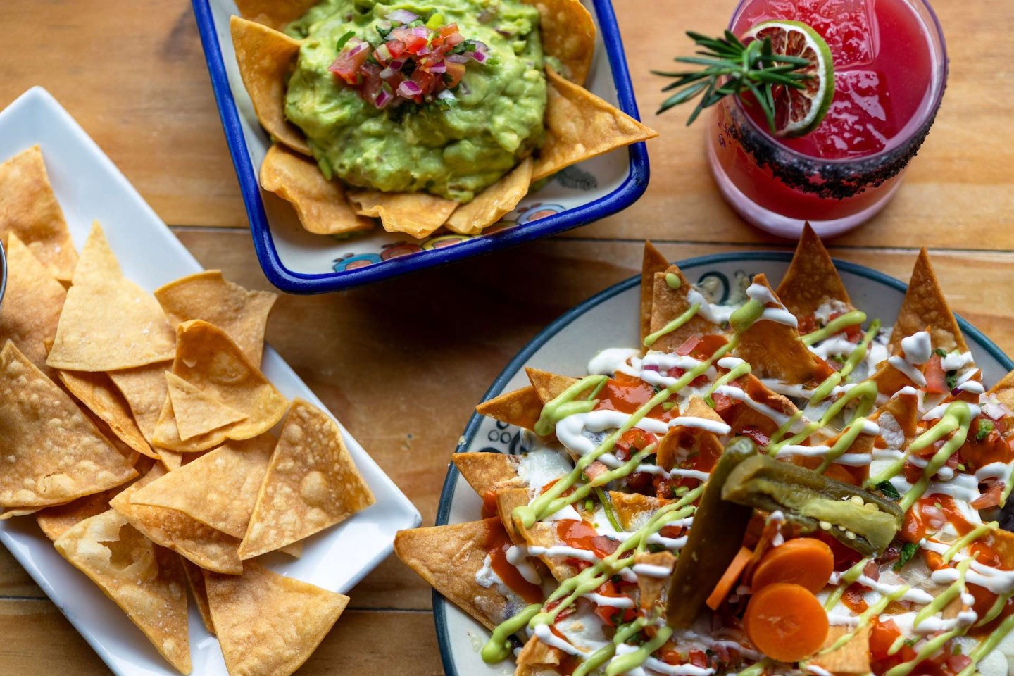 Colorful Mexican meal featuring nachos topped with sauces and pickled vegetables, a bowl of guacamole with tortilla chips, extra chips on a plate, and a pink drink garnished with a sprig of rosemary on a wooden table.
