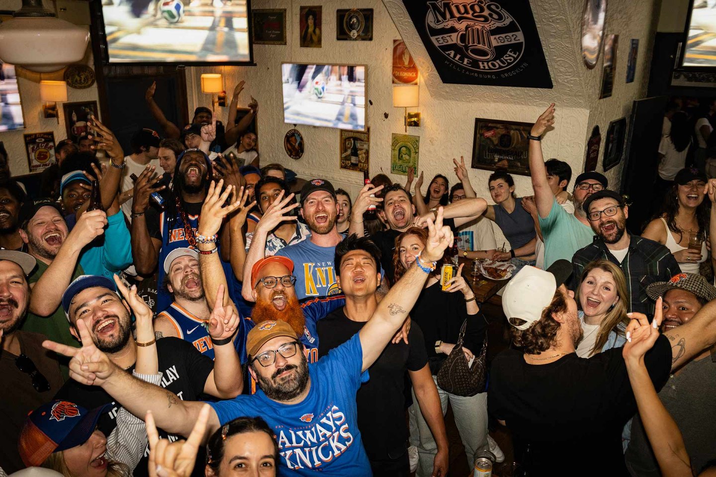 A lively crowd of sports fans, many wearing Knicks gear, cheer and raise their hands inside a bar decorated with sports memorabilia and multiple TVs showing a game.