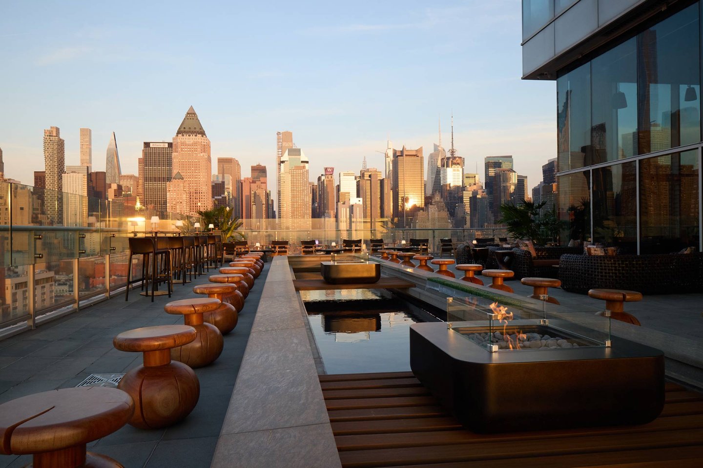 A rooftop bar with wooden stools and tables, sleek fire pits, and glass railings overlooks the New York City skyline at sunset. Tall buildings and skyscrapers are visible in the background.