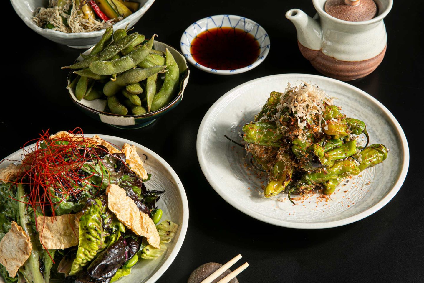 A selection of Japanese dishes on a black table, including a salad with greens and crispy toppings, a bowl of edamame, grilled shishito peppers with bonito flakes, dipping sauce, and a teapot.