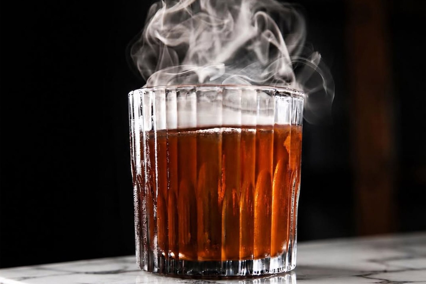 A glass of dark brown cocktail emits wisps of smoke, sitting on a white marble surface against a dark background. The drink is served in a faceted, clear glass.