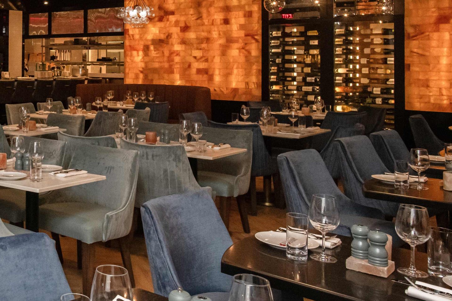 Restaurant interior with tables set for dining, featuring blue and gray chairs. Walls display warm wooden panels and a wine rack. The ambiance is elegant, with neatly arranged glassware and cutlery on the tables. Hanging lights add a soft glow.