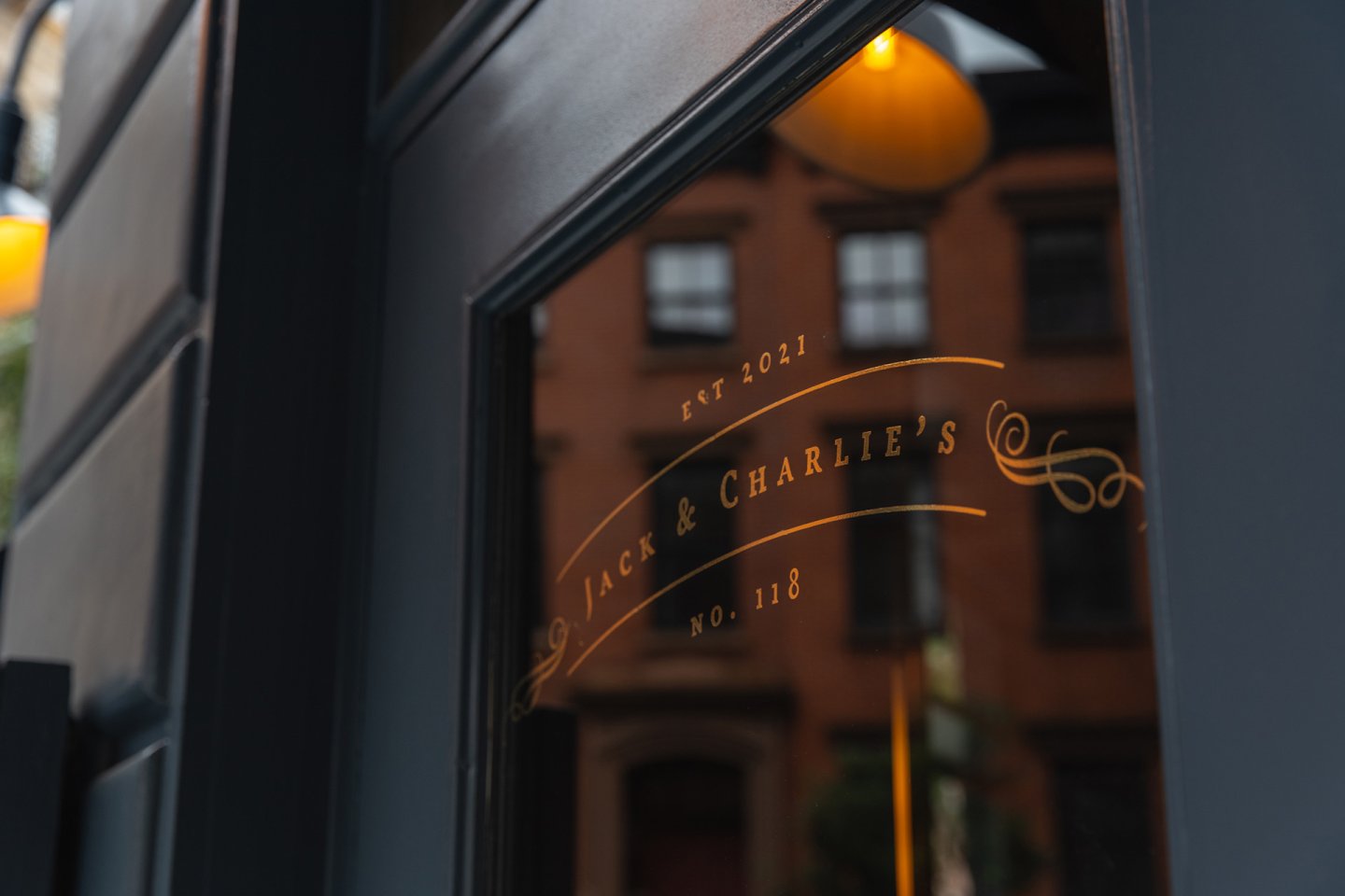 A glass door with gold lettering that reads "Est 2021 Jack & Charlie’s No. 118" reflects the facade of a brick building across the street.