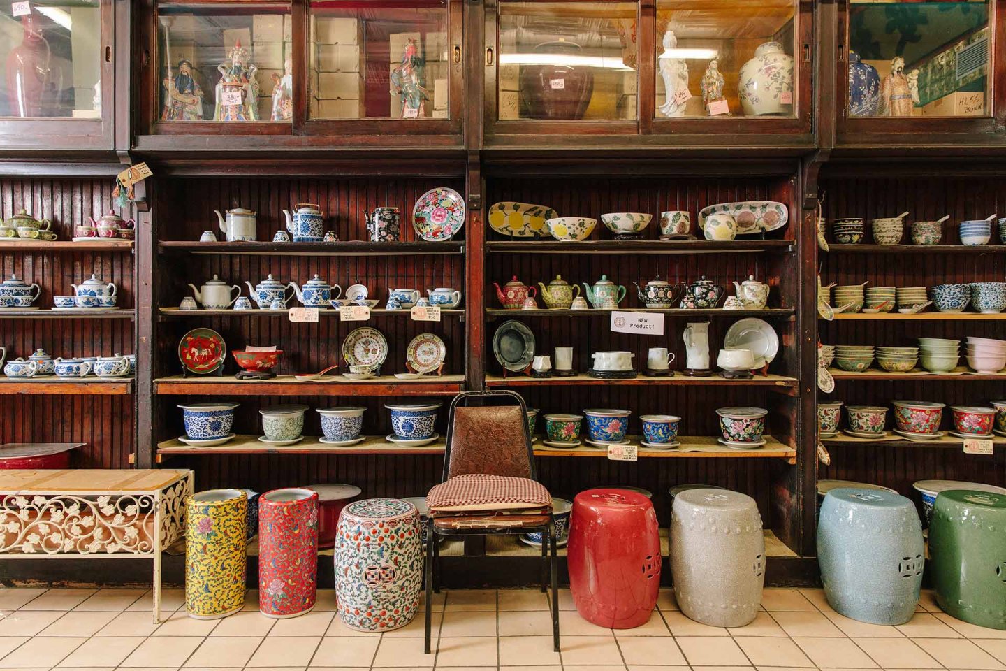 Shelves filled with colorful ceramic bowls, plates, and teapots line a wooden wall. Patterned stools and chairs are arranged in front, and various decorative items are displayed above the shelves.