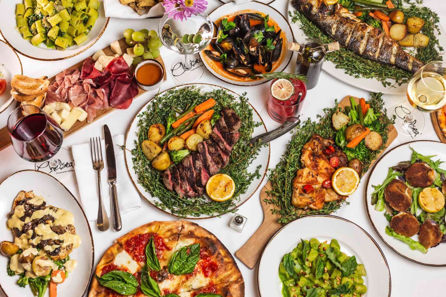 A lavish spread of Italian cuisine featuring dishes like pizza, steak with vegetables, grilled fish, and assorted cheeses. Accompanied by wine and garnished with fresh herbs, the table displays a vibrant and appetizing array of meals.