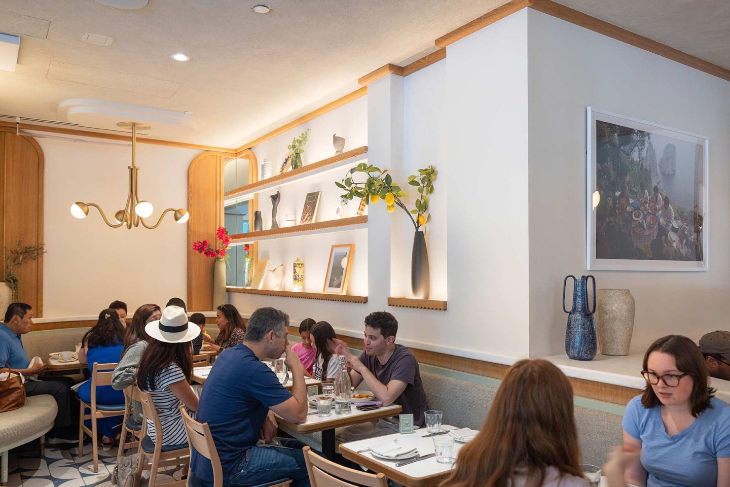 People sit at tables in a modern, well-lit restaurant with white walls, wooden accents, shelves with decor, and natural light. Some are eating or talking, creating a lively and relaxed atmosphere.