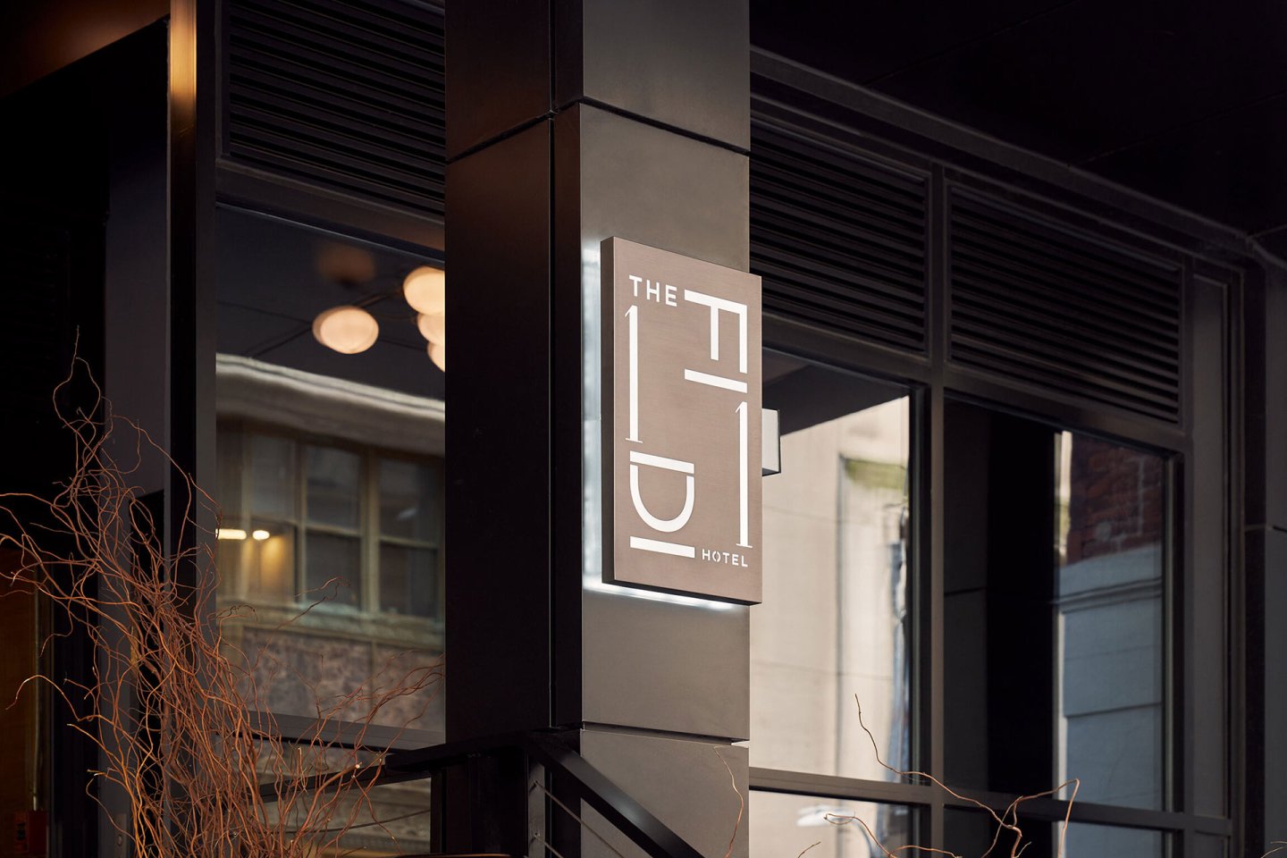 A modern hotel sign reading "The FIDI Hotel" is mounted on a dark exterior wall. Reflections of nearby buildings and ceiling lights are visible in the large windows behind the sign.