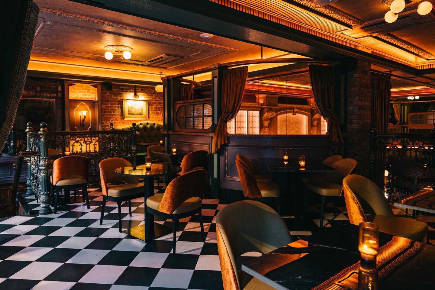 Dimly lit lounge with checkered floors, velvet chairs, dark tables, warm lighting, and curtained windows create a cozy, elegant atmosphere. Candles add a soft glow throughout the space.