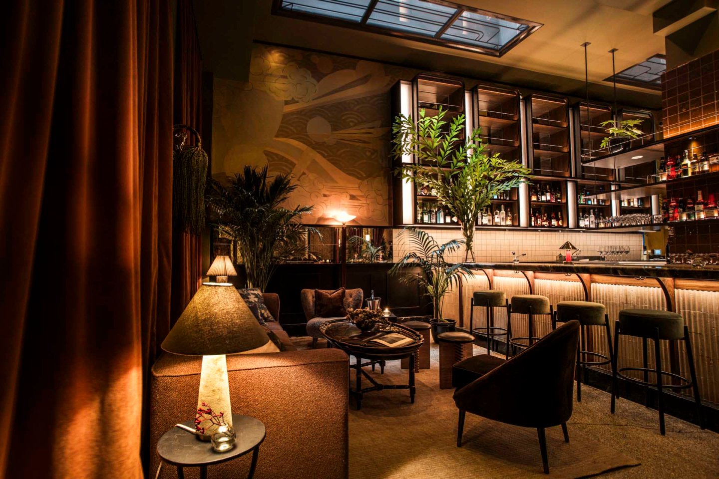 A dimly lit, cozy bar with a warm ambiance. It features a plush seating area with a small table lamp, high stools at the bar, shelves lined with bottles, and decorative plants. The ceiling has skylights, and there are rich curtains on one side.