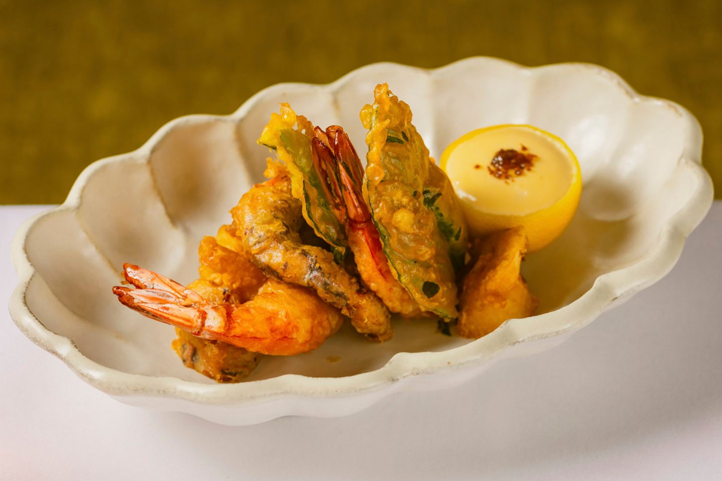 A scalloped white dish holds several pieces of crispy fried shrimp and vegetables, garnished with a lemon half filled with dipping sauce. The dish is placed on a white surface against a green background.