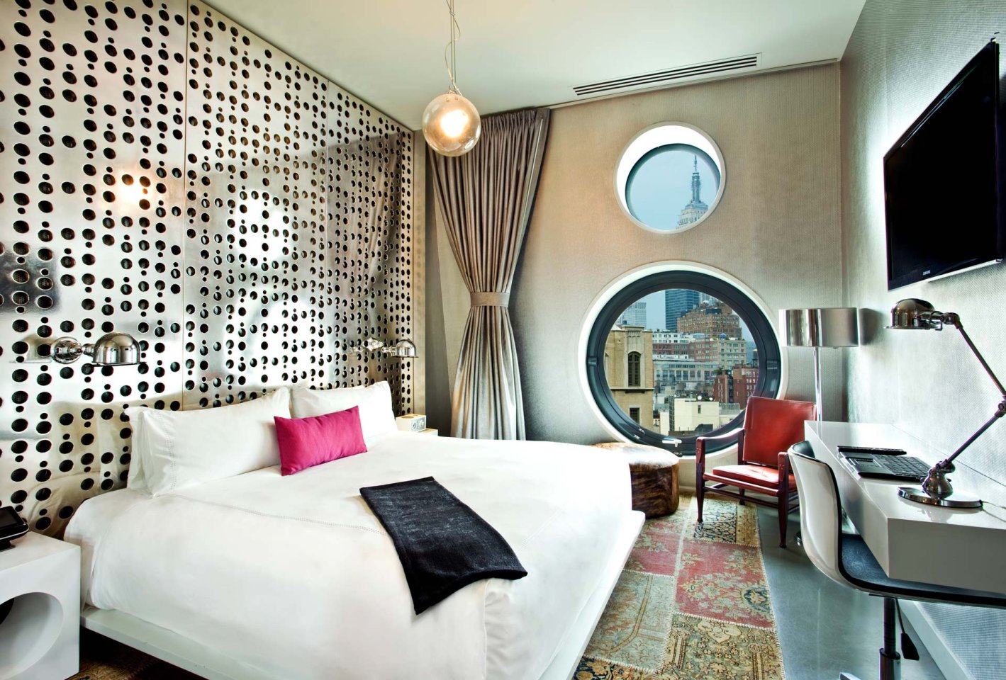 Modern hotel room with a large bed, white bedding, pink accent pillow, patterned accent wall, two round windows with city views, desk with chair, wall-mounted TV, and contemporary decor.