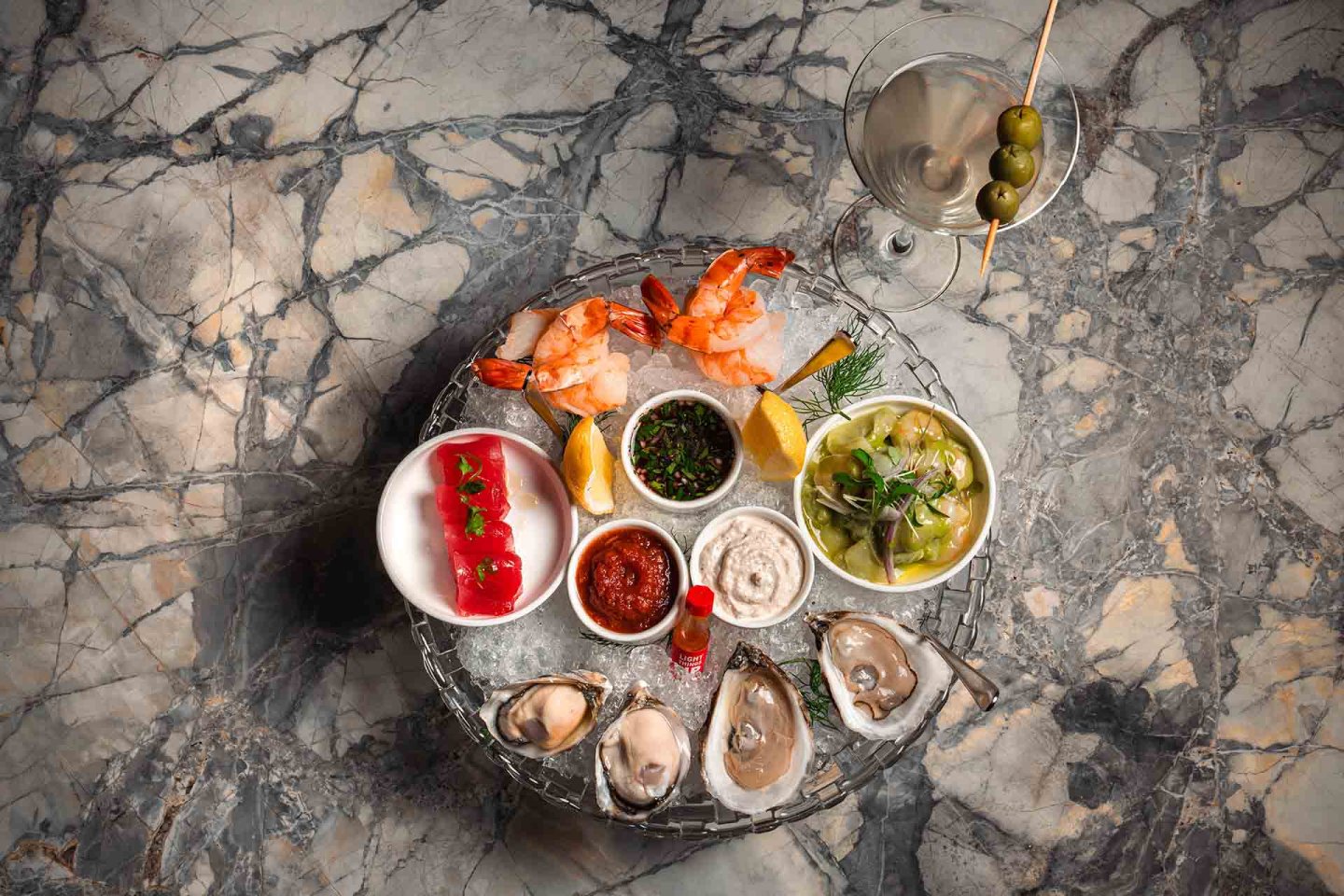 A marble table holds a seafood platter with oysters, shrimp, lemon wedges, sauces, tuna tartare, ceviche, and a martini garnished with green olives.