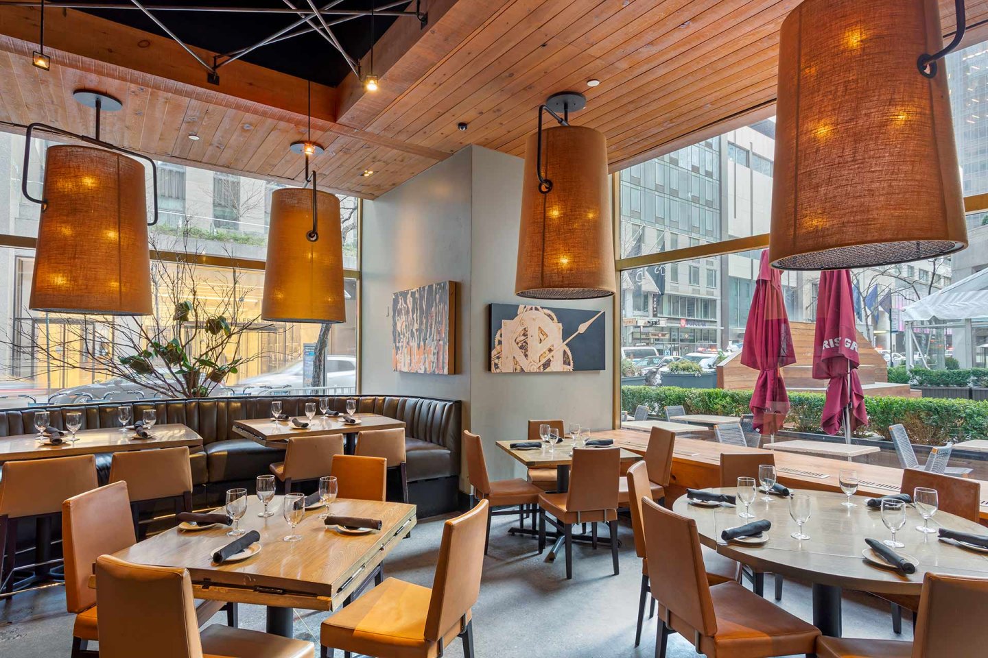 Modern restaurant interior with large hanging lamps, wooden tables set with glasses and napkins, cushioned booth seating, and large windows showing city buildings and outdoor patio with red umbrellas.