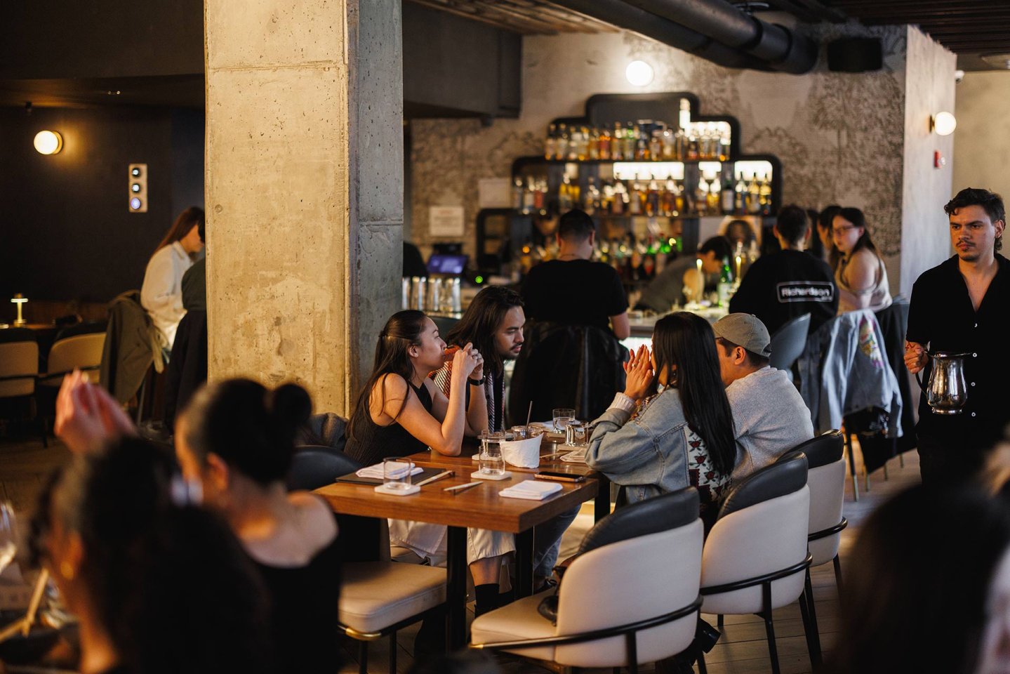 A lively restaurant scene with people sitting at tables, eating, drinking, and chatting. A waiter with a coffee pot walks by. The bar in the background is stocked with bottles, and the atmosphere appears warm and social.