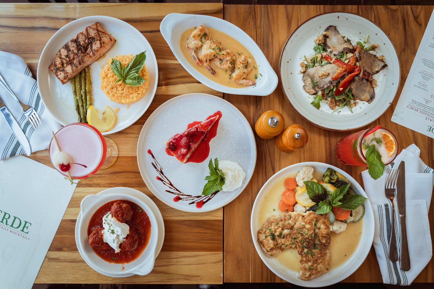 A wooden table set with plates of various dishes, including grilled salmon, meatballs, roasted meats, vegetables, a dessert with berries and cream, cocktails, and a menu, viewed from above.