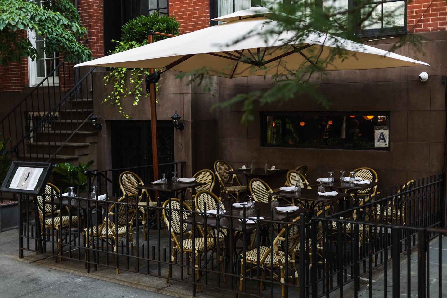 Outdoor patio of a restaurant with empty woven chairs and tables, surrounded by a black metal fence, under a large white umbrella, next to a brick building.
