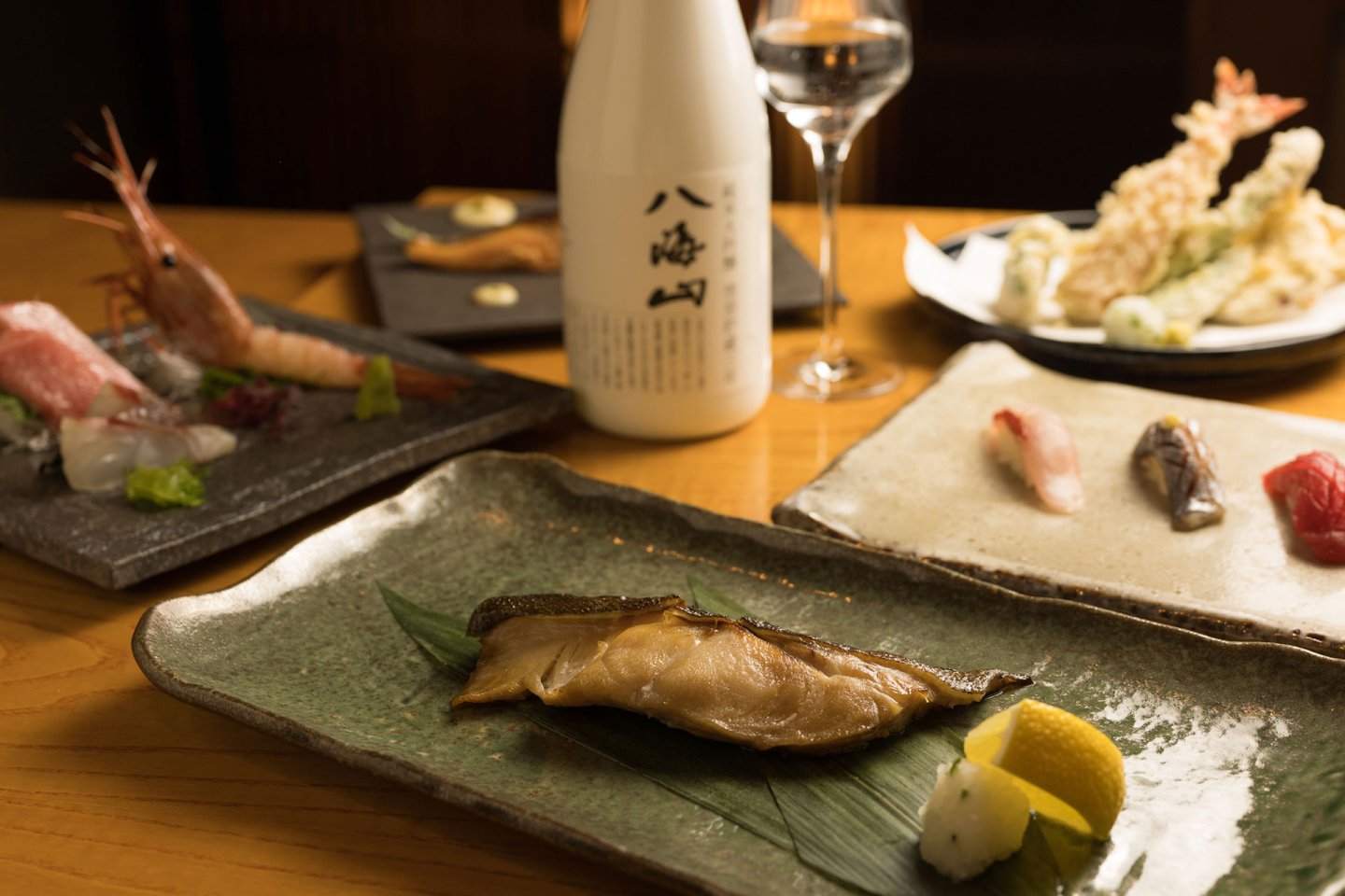 A Japanese meal with grilled fish on a green plate, assorted sashimi, tempura, and sushi, served with a bottle of sake and a glass on a wooden table.