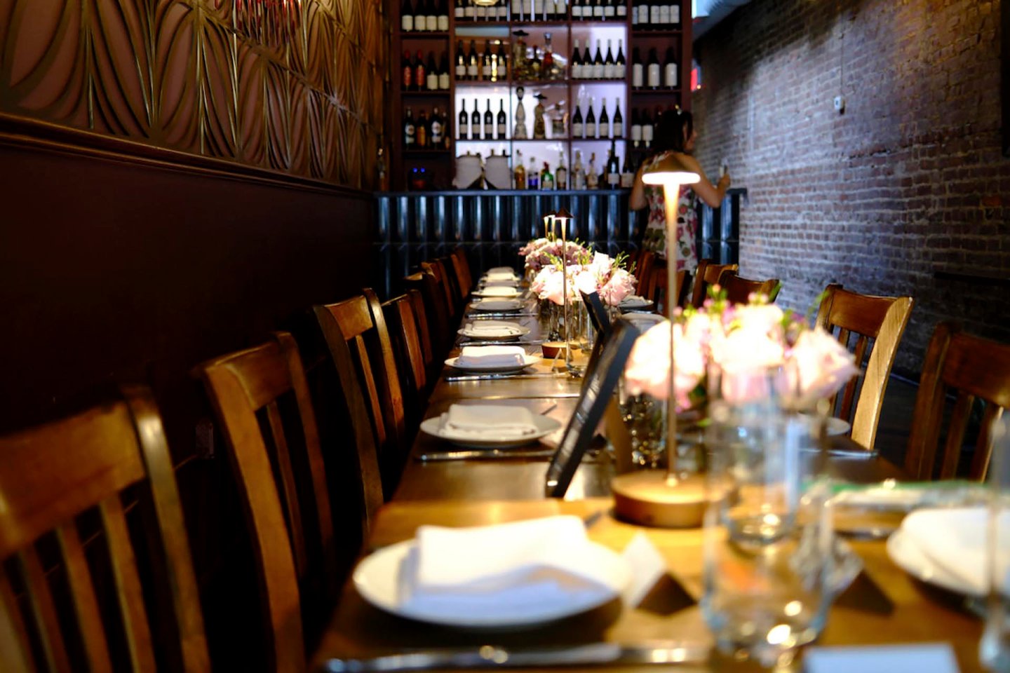 A long wooden dining table set for a meal with white plates, glasses, and floral centerpieces. The restaurant has dim lighting, a brick wall on the right, and a shelf filled with wine bottles in the background.