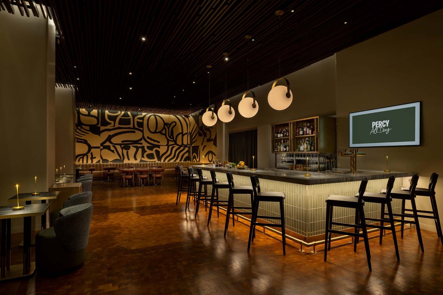 A modern bar with a geometric mural, stylish lighting, a tiled bar counter, black barstools, and a wall-mounted screen reading "PERCY All Day." Tables with candles and lounge chairs are on the left. The atmosphere is warm and inviting.