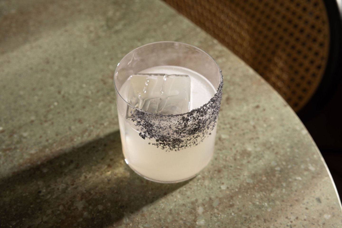 A clear cocktail with a large ice cube is served in a glass with a black salt rim, placed on a green speckled table next to a woven chair.