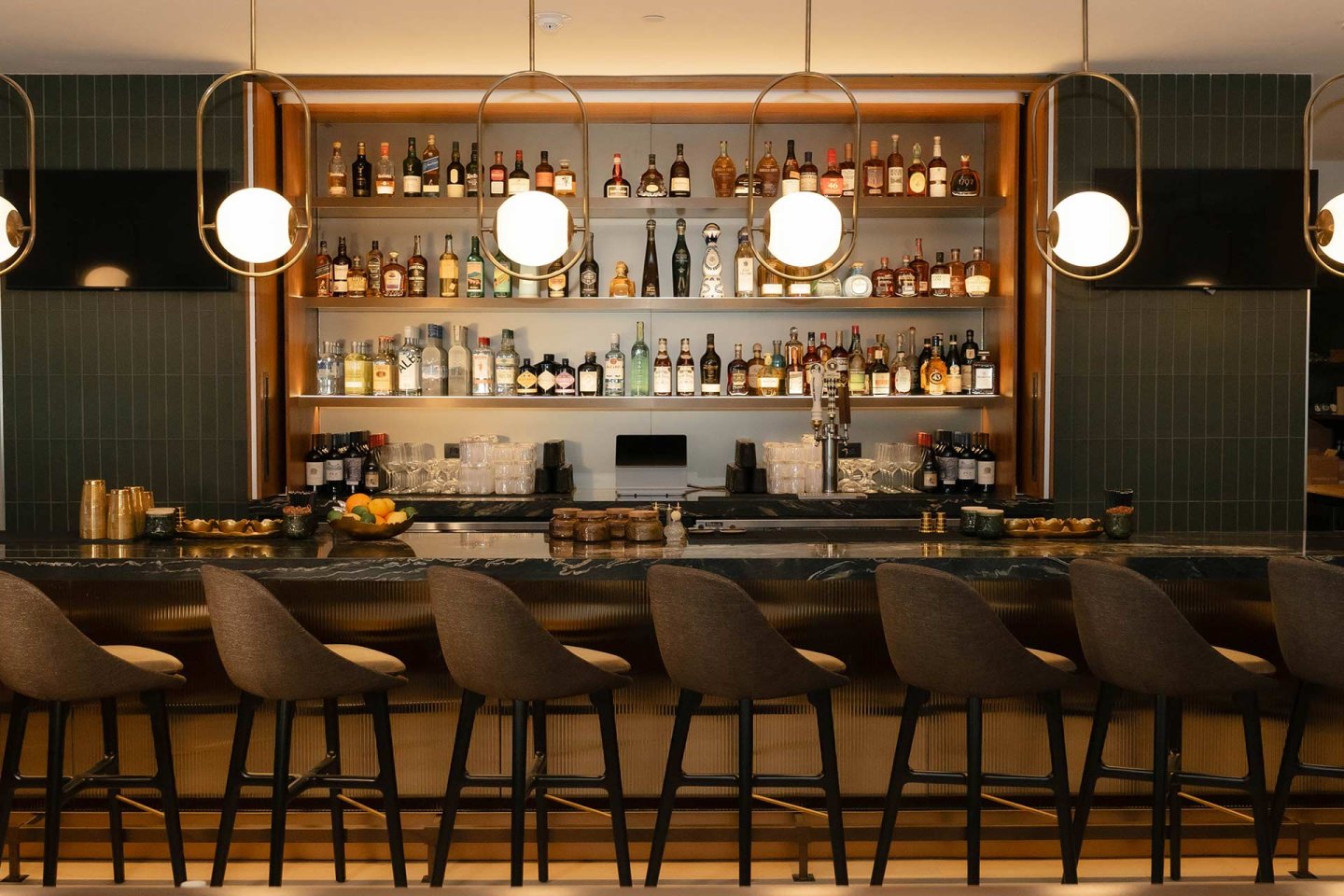 A modern bar with high-backed chairs, globe pendant lights, and shelves stocked with assorted liquor bottles behind a long, marble countertop.