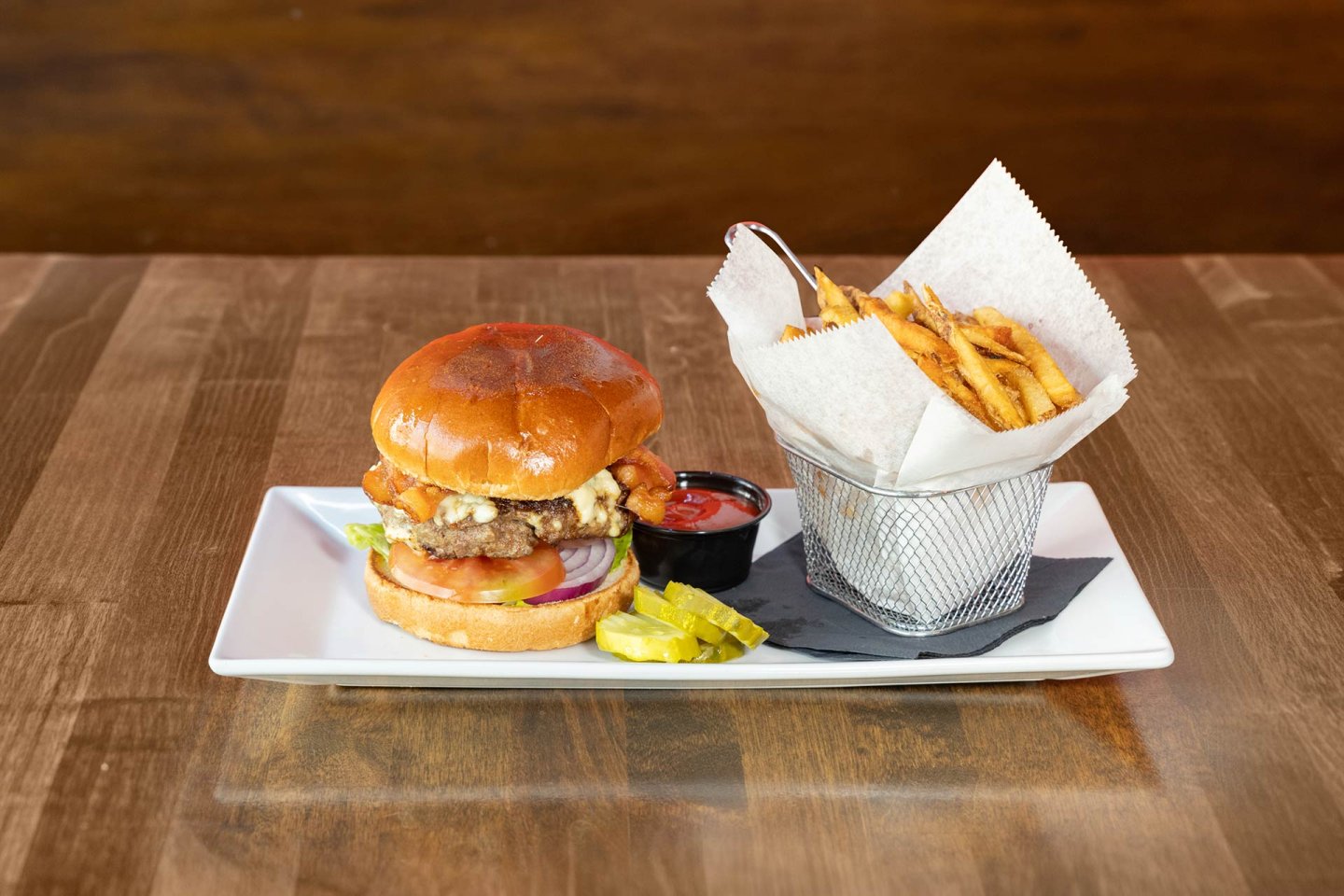 A cheeseburger with lettuce, tomato, onion, and pickles sits on a white plate next to a basket of French fries and a small cup of ketchup on a wooden table.