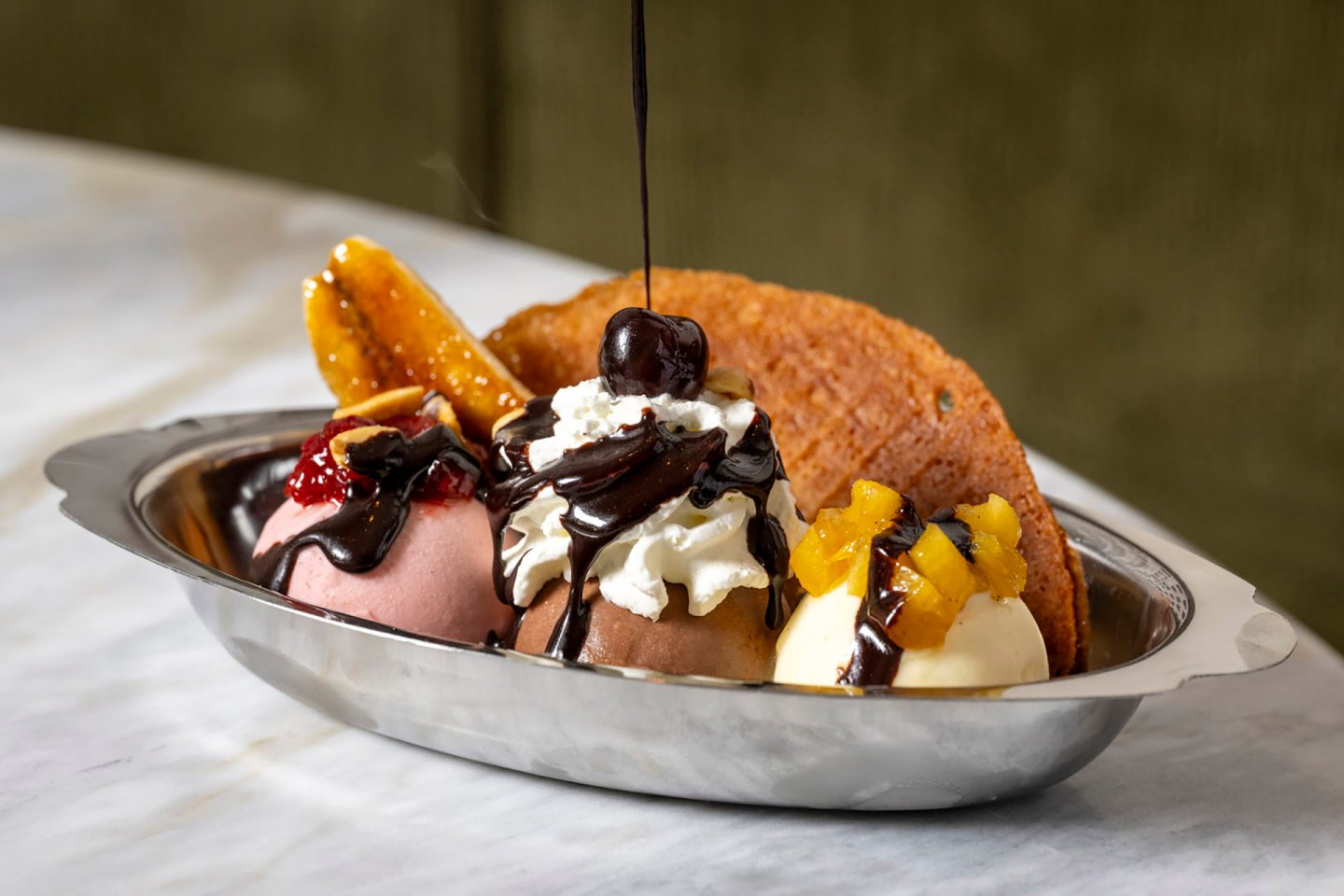 A banana split served in a silver dish with three scoops of ice cream topped with chocolate sauce, whipped cream, a cherry, and fruit garnishes. A waffle slice is placed on the side.