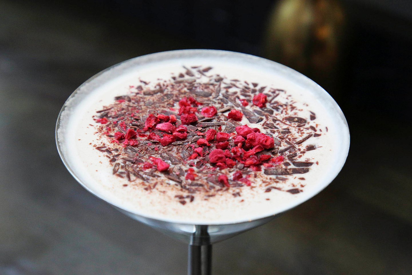 A creamy cocktail in a martini glass, topped with shaved chocolate and crushed freeze-dried raspberries. The drink is set on a dark, blurred background.