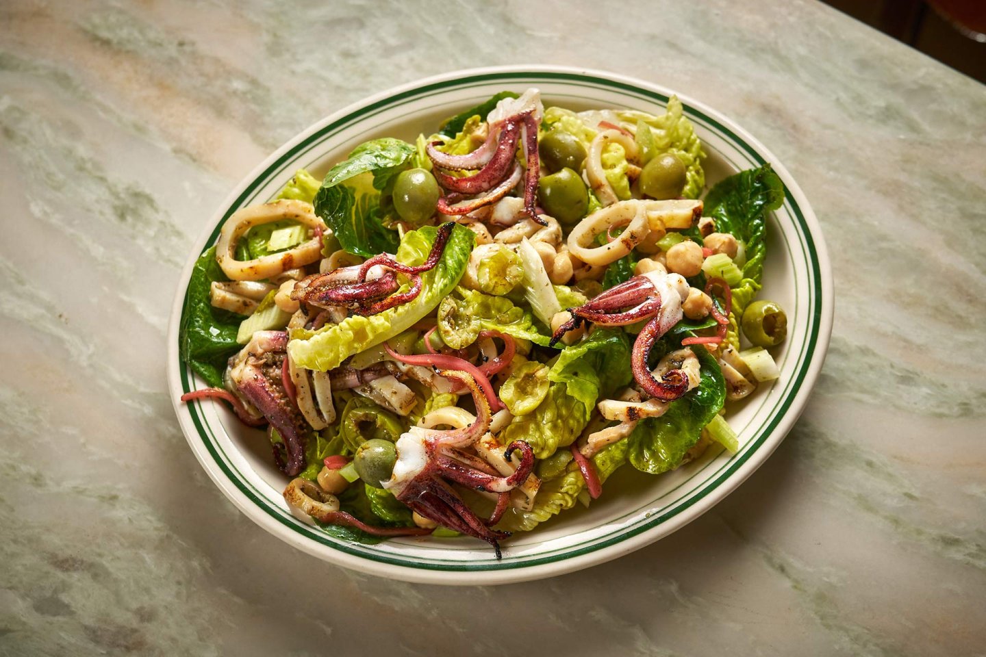 A plate of fresh salad with leafy greens, green olives, chickpeas, and pieces of grilled squid and octopus, served on a white plate with green trim on a marble surface.