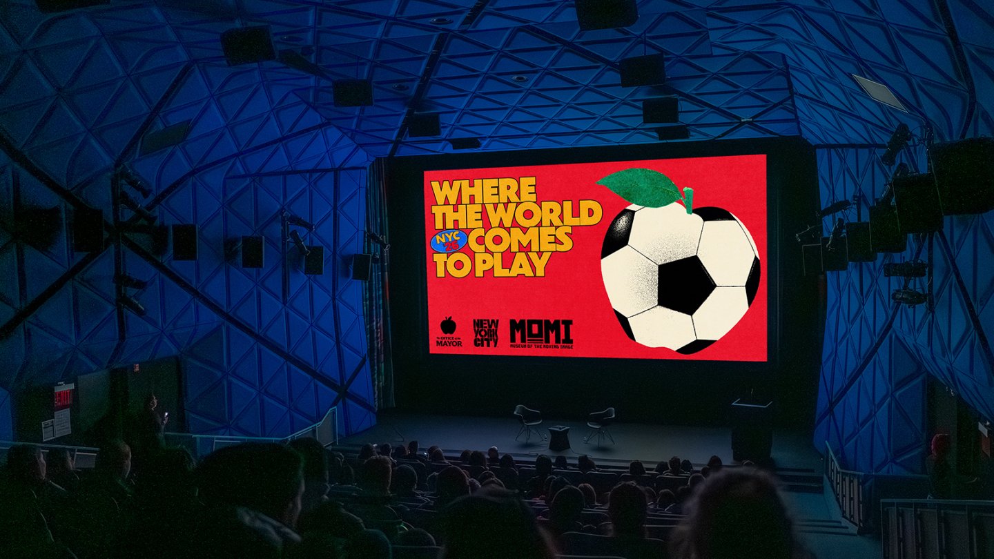a scene of a theater with blue felt walls and ceiling with people in the seats and a bright image on the screen that is a promotional graphic for "Where the World Comes to Play" showing a soccer ball in the shape on an apple.
