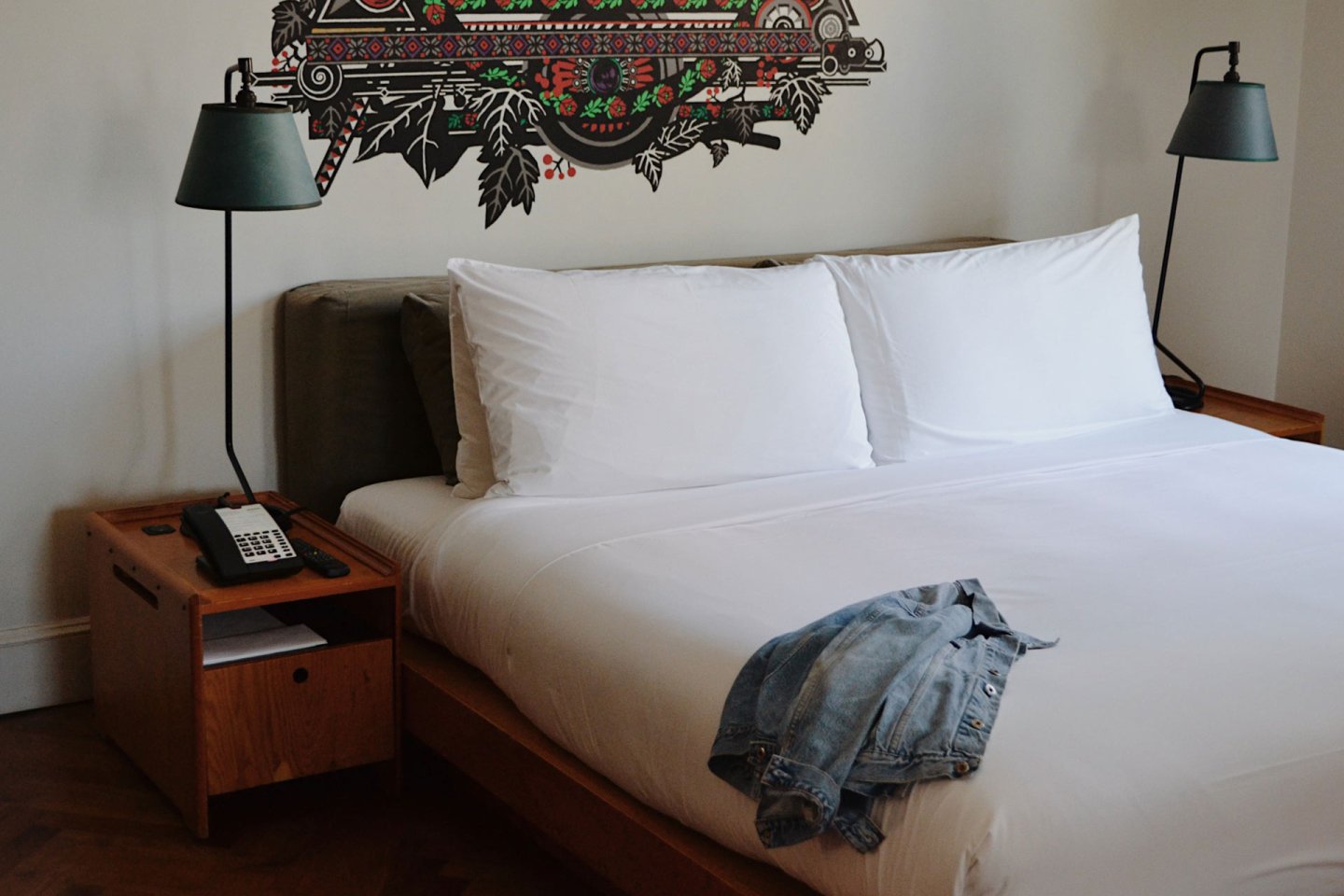 A neatly made bed with white linens and two pillows, flanked by wooden nightstands with lamps. A phone sits on the left nightstand, and a denim jacket is draped across the end of the bed.