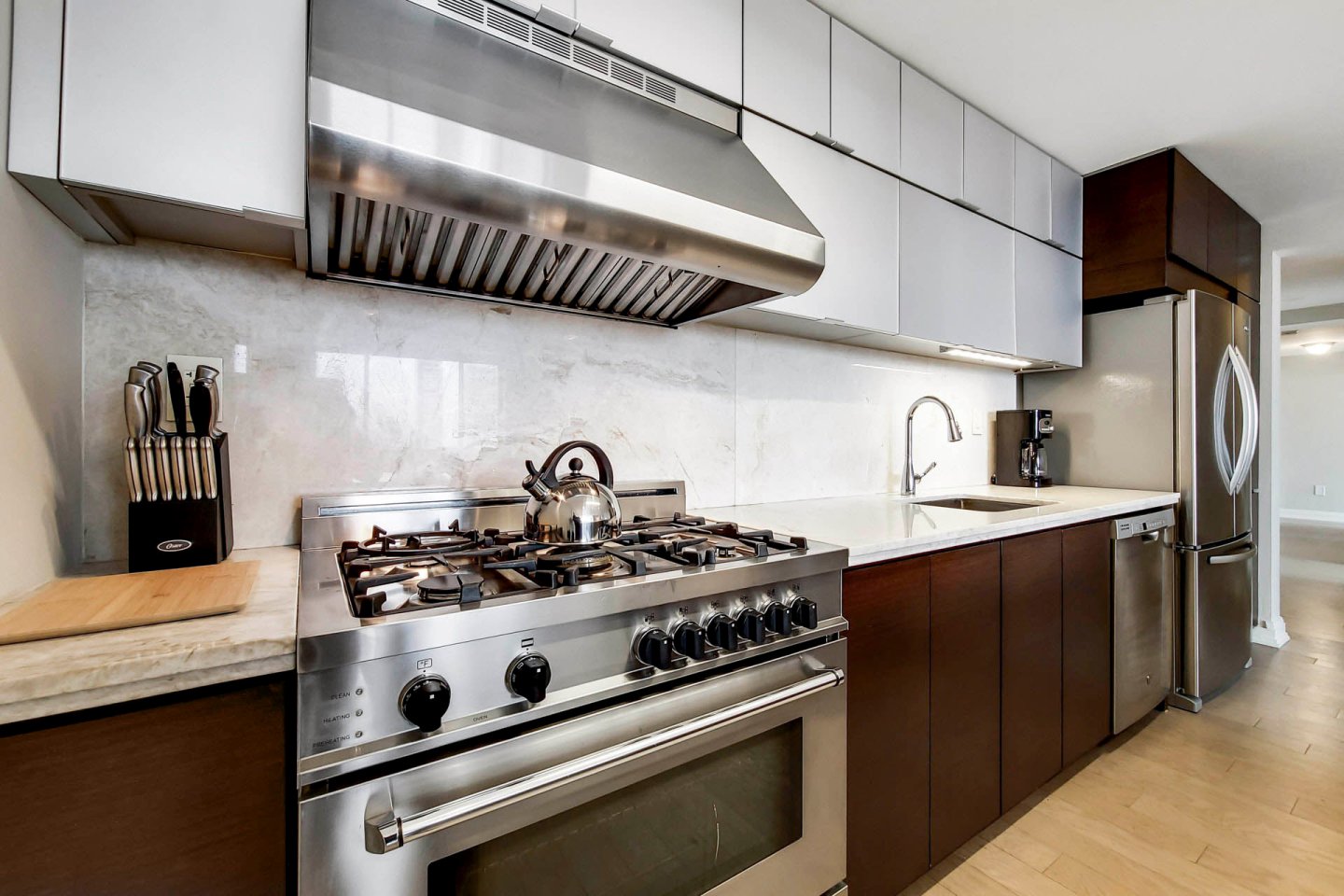 Modern kitchen with stainless steel appliances, including a large gas stove and range hood. A silver kettle sits on the stove. White and dark wood cabinets line the walls, and a set of knives rests on a wooden block. Light-colored flooring throughout.