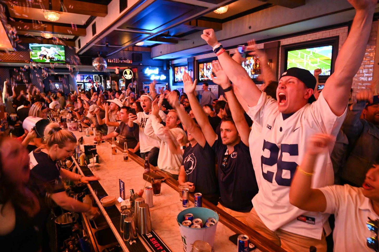 A lively crowd at a sports bar cheers and raises their arms in excitement while watching a football game on TVs. People wear sports jerseys, and drinks are spread across the bar. The atmosphere is energetic and festive.