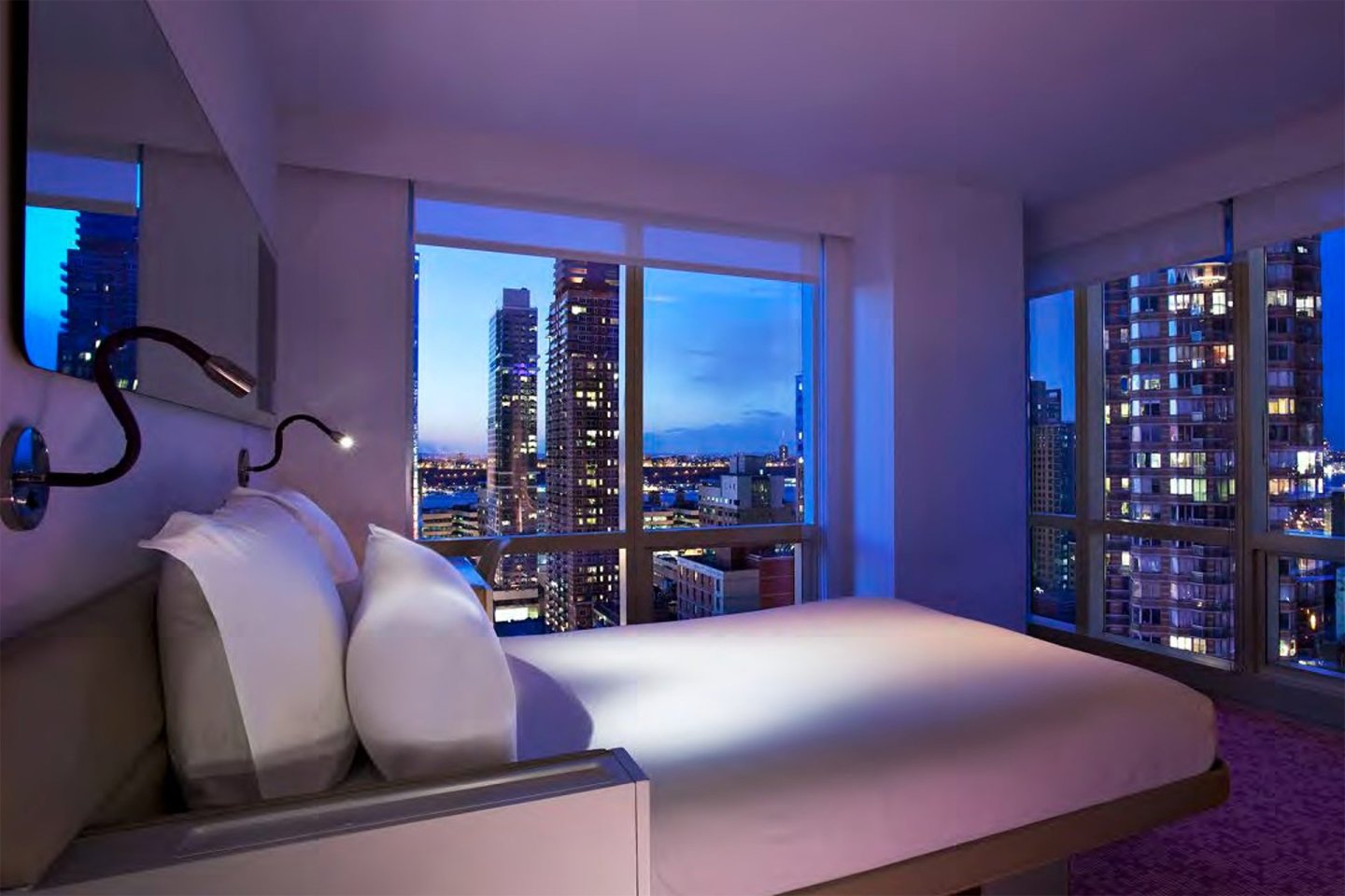 A modern hotel room with large windows offers a night view of a city skyline with tall buildings and lights. The room features a neatly made bed with white linens and a reading lamp.
