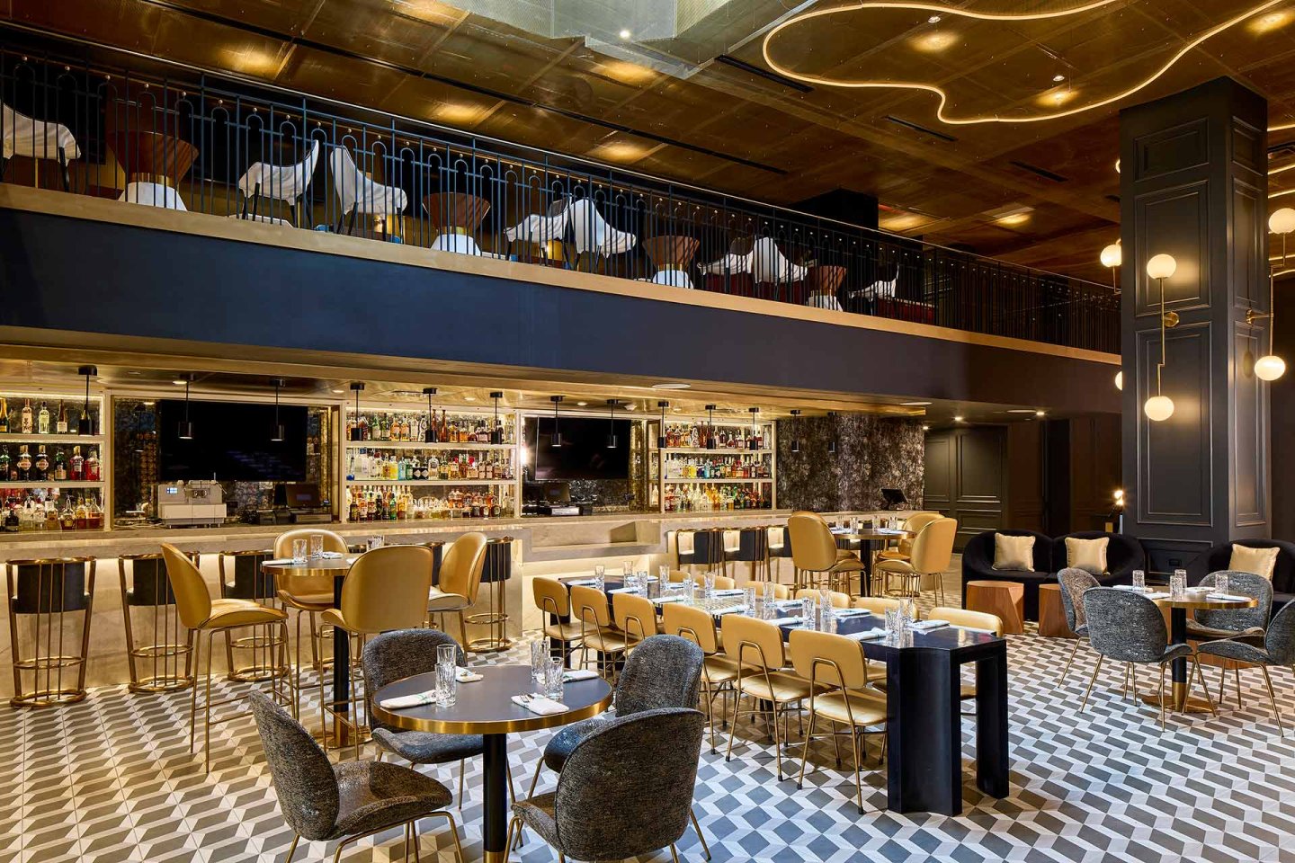Modern, upscale restaurant interior with geometric tile flooring, gold and gray furnishings, a long bar with bottles on display, and an upper mezzanine level with additional seating. Elegant lighting fixtures hang from a gold ceiling.