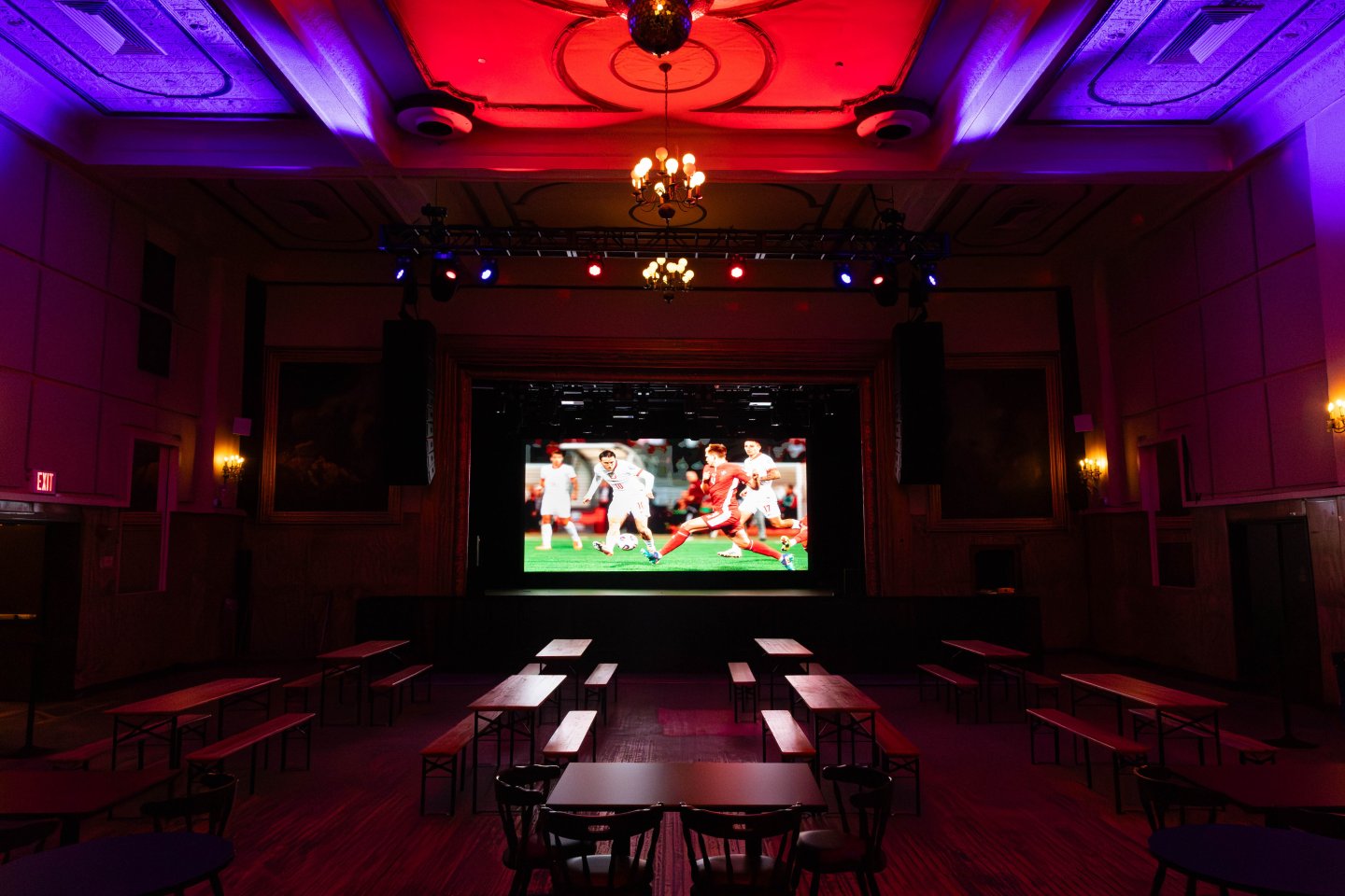 Photo of Warsaw's main hall featuring 12 large empty picnic tables in front of a 24 foot video wall showing professional soccer placers kicking a ball during a soccer game.